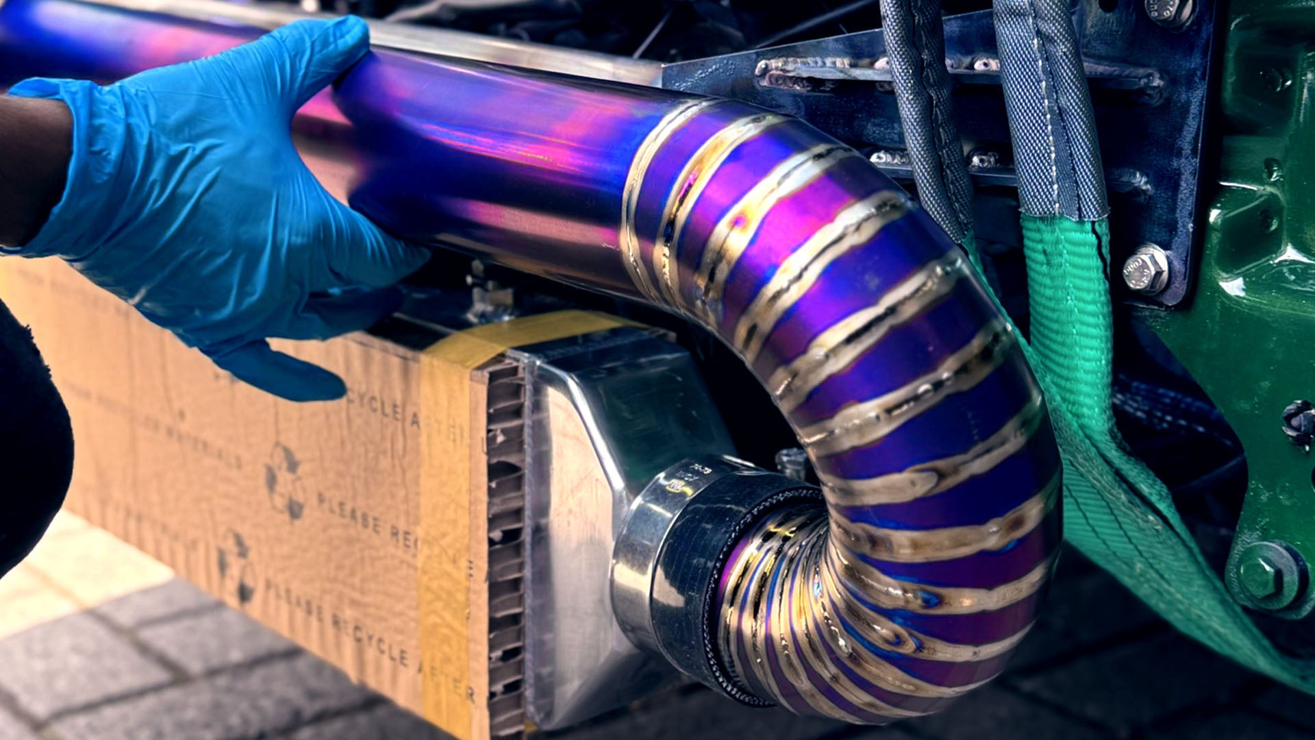Monteur met blauwe handschoen monteert een custom TIG-gelaste titanium intercoolerbuis met paarse hitteverkleuring op een aluminium intercooler.