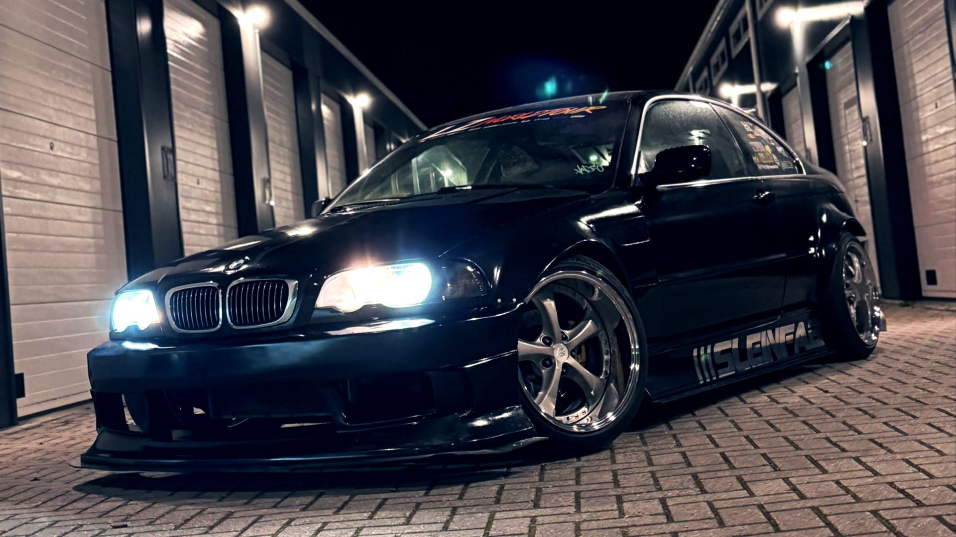 Zwarte getunede BMW E46 coupé met brandende koplampen en SleinFab sticker 's nachts geparkeerd voor verlichte garageboxen.